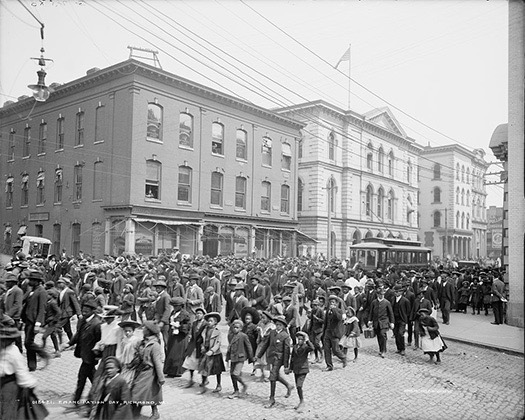 Black-and-white photograph of a large street parade with families, children, and public gathering lines.