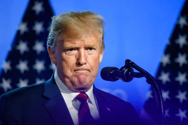 President Donald Trump standing at a microphone against a blue backdrop with American flags.